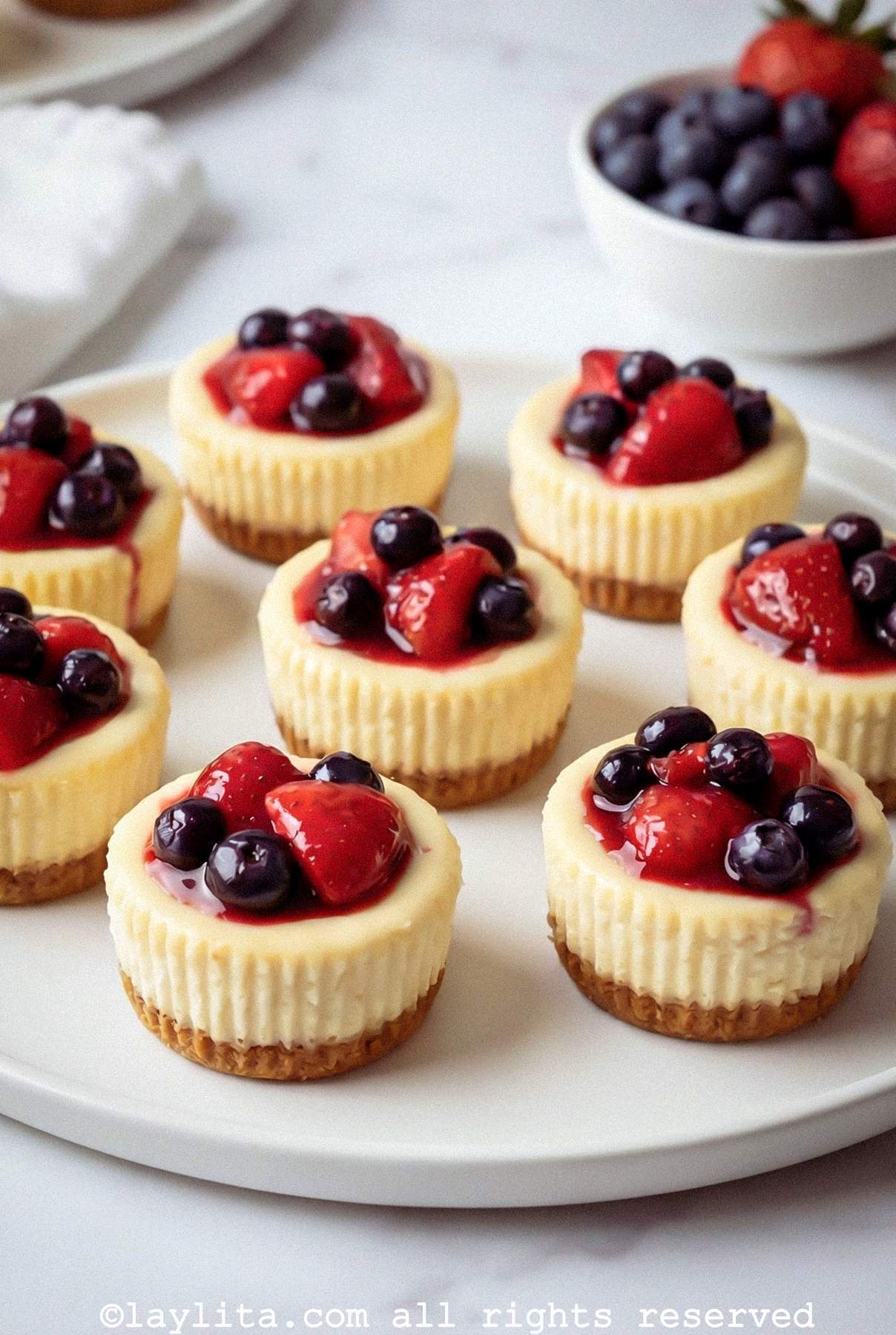 Mini cheesecakes topped with mixed berries on a white platter with more berries in the background
