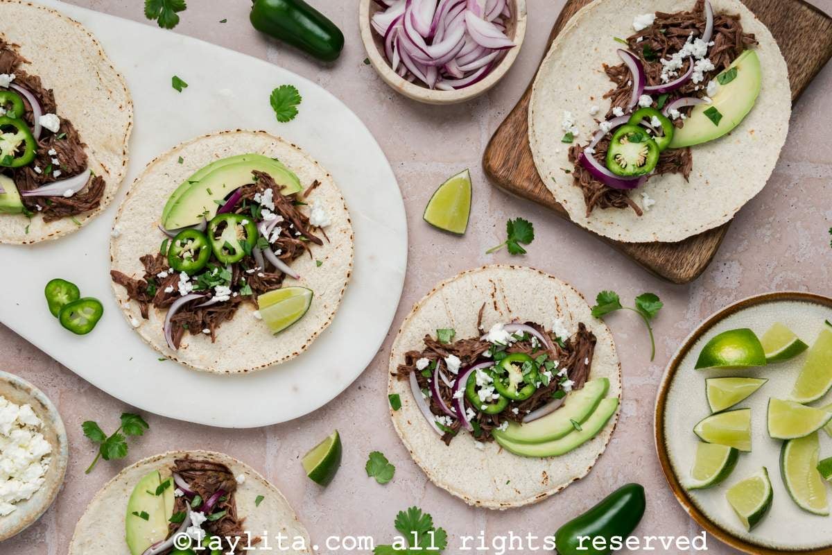 Tablescape of barbacoa tacos, with some on plates and others on the counter or a wooden board.