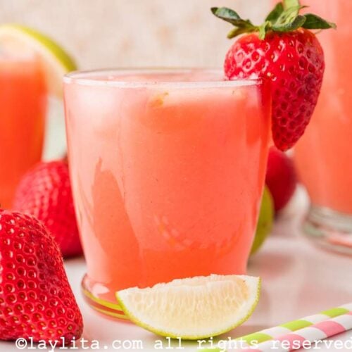 Side view of a glass of strawberry agua fresca with a lime wedge and a whole strawberry in front of it.