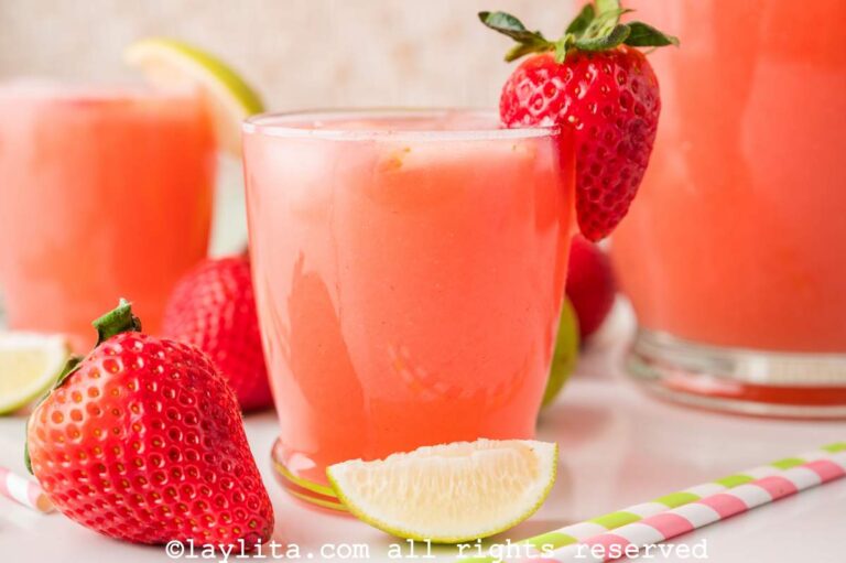 Side view of a glass of strawberry agua fresca with a lime wedge and a whole strawberry in front of it.