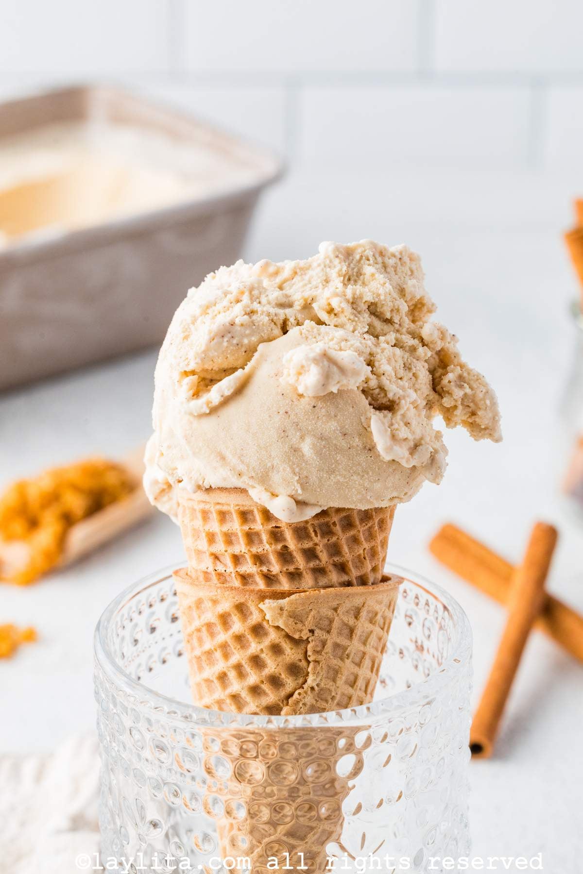 A double waffle cone filled with a large scoop of cinnamon ice cream in a glass cup, with cinnamon sticks in the background