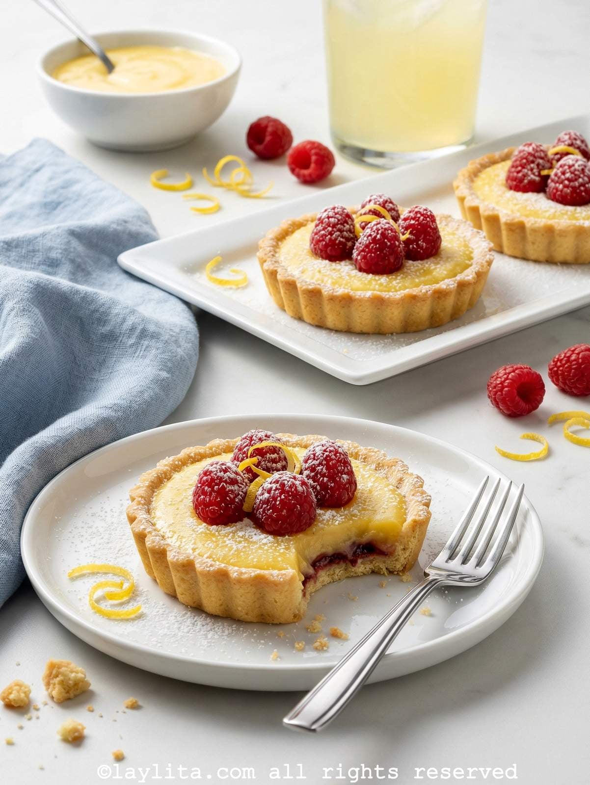 Raspberry lemon tartlet served on a white plate with one bite missing from the tart. In the background there are more tartlets on a platter