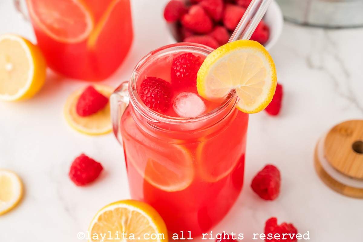 A glass of raspberry lemonade garnished with lemon slices on a counter with raspberries and lemons spread around, in the background there is another glass of lemonade