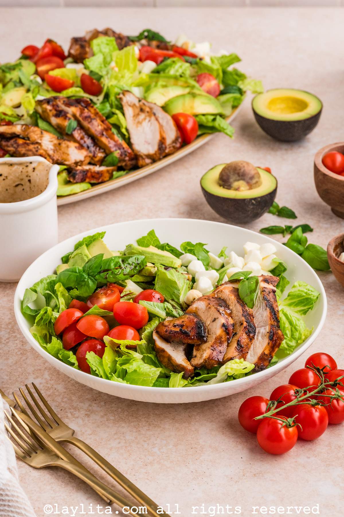 A white bowl filled with lettuce salad and topped with mozzarella, tomatoes, avocado slices and grilled chicken. In the background there is a platter with more salad on it.