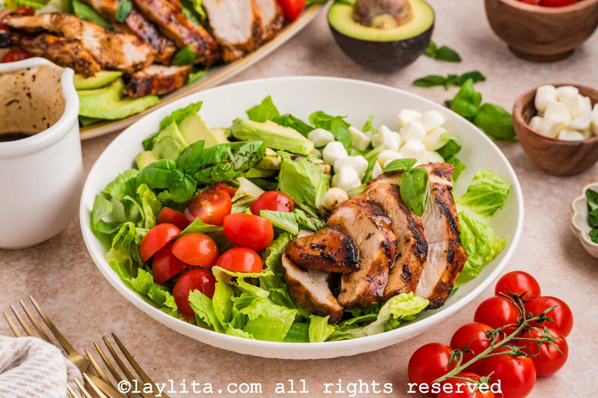 A white bowl filled with lettuce salad and topped with mozzarella, tomatoes, avocado slices and grilled chicken. In the background there is a platter with more salad on it.
