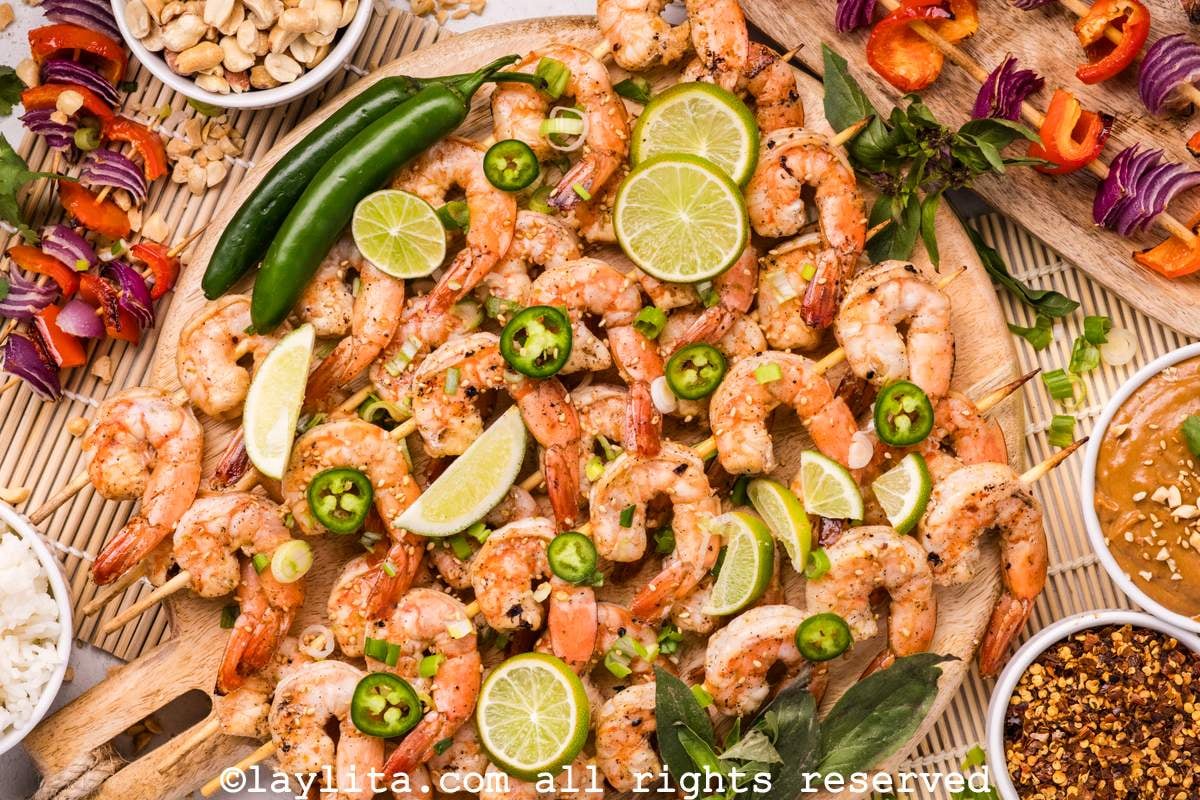 Overhead shot of a platter of grilled shrimp skewers with spicy peppers, veggie skewers, chili flakes and peanut sauce on the side