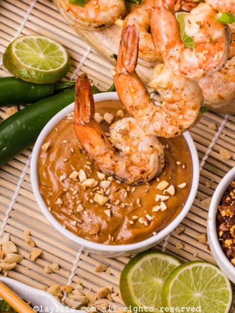 A shrimp skewer being dipped into a white ramekin filled with peanut sauce, on the side there are spicy peppers and lime slices