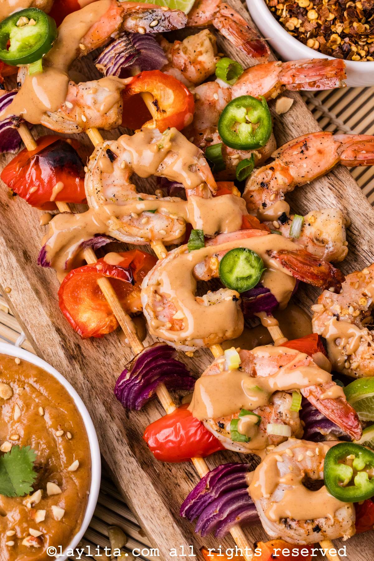 View of several shrimp skewers drizzled with peanut sauce and topped with serrano pepper slices, with a bowl of peanut sauce and a veggie skewer on the side
