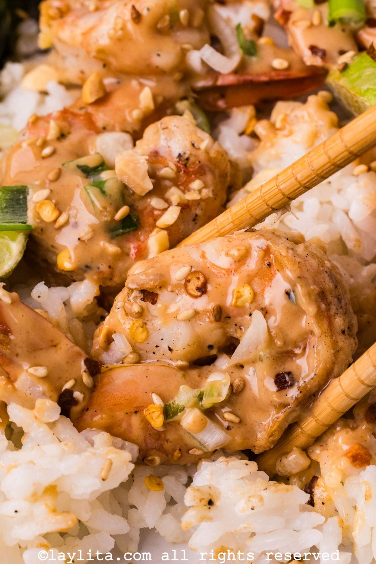 A set of chopsticks holding a shrimp with peanut sauce on a bed of rice, with more shrimp on the side