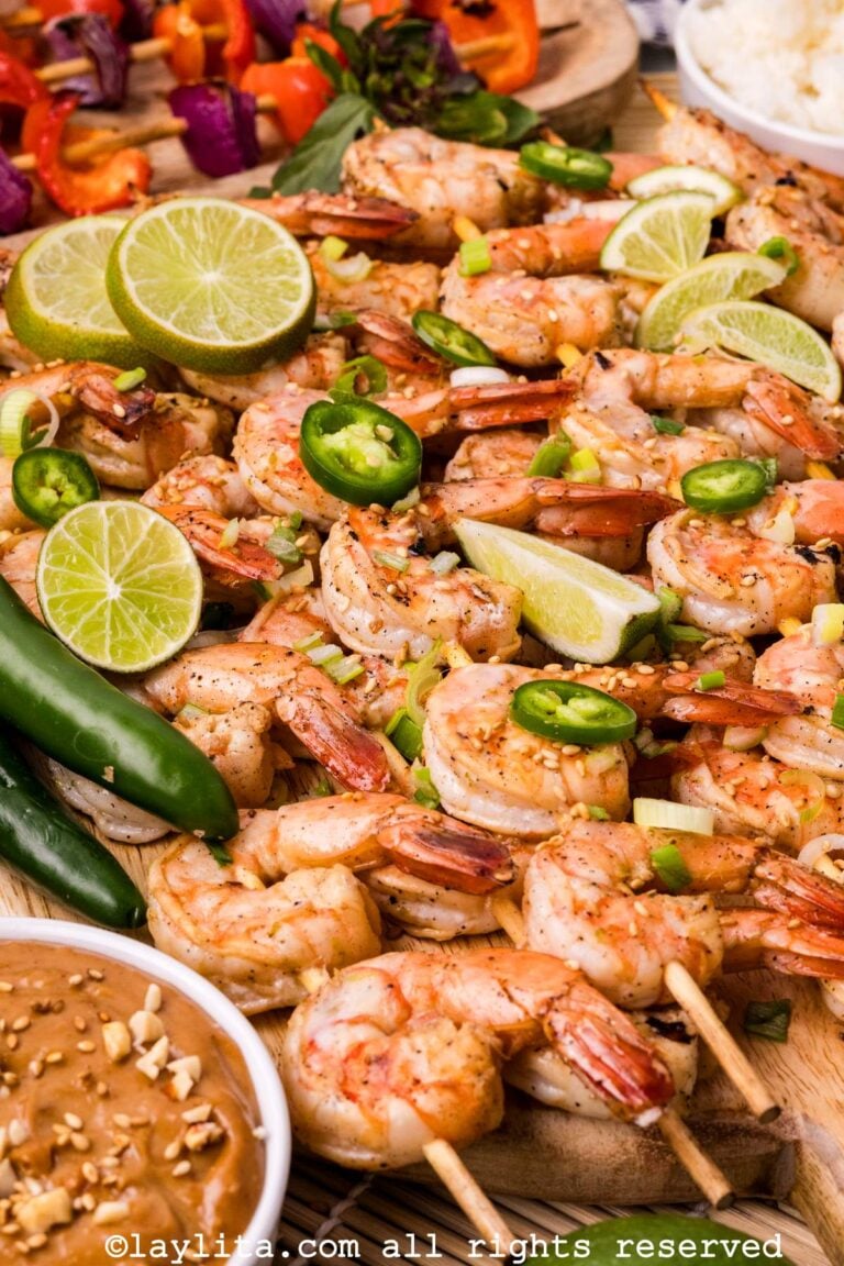 Side view of several shrimp skewers topped with sliced serrano peppers, lime slices, green onions, next to a bowl of peanut sauce