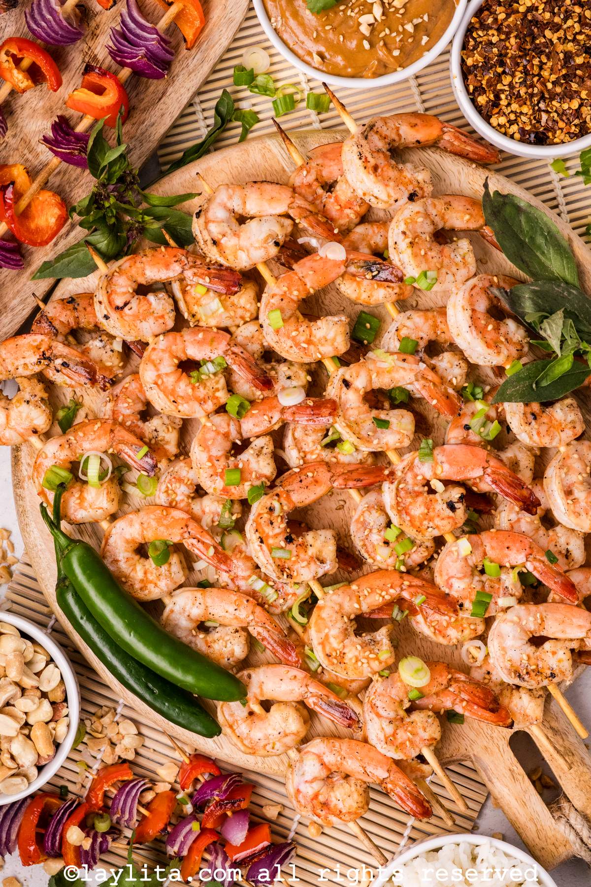 Overhead shot of a platter of grilled shrimp skewers with spicy peppers, veggie skewers, chili flakes and peanut sauce on the side