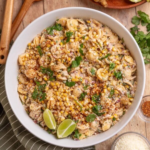 Overhead shot of a bowl filled with Mexican style corn pasta salad next to a tray of grilled corn, some wooden spoons and a bowl of cotija cheese