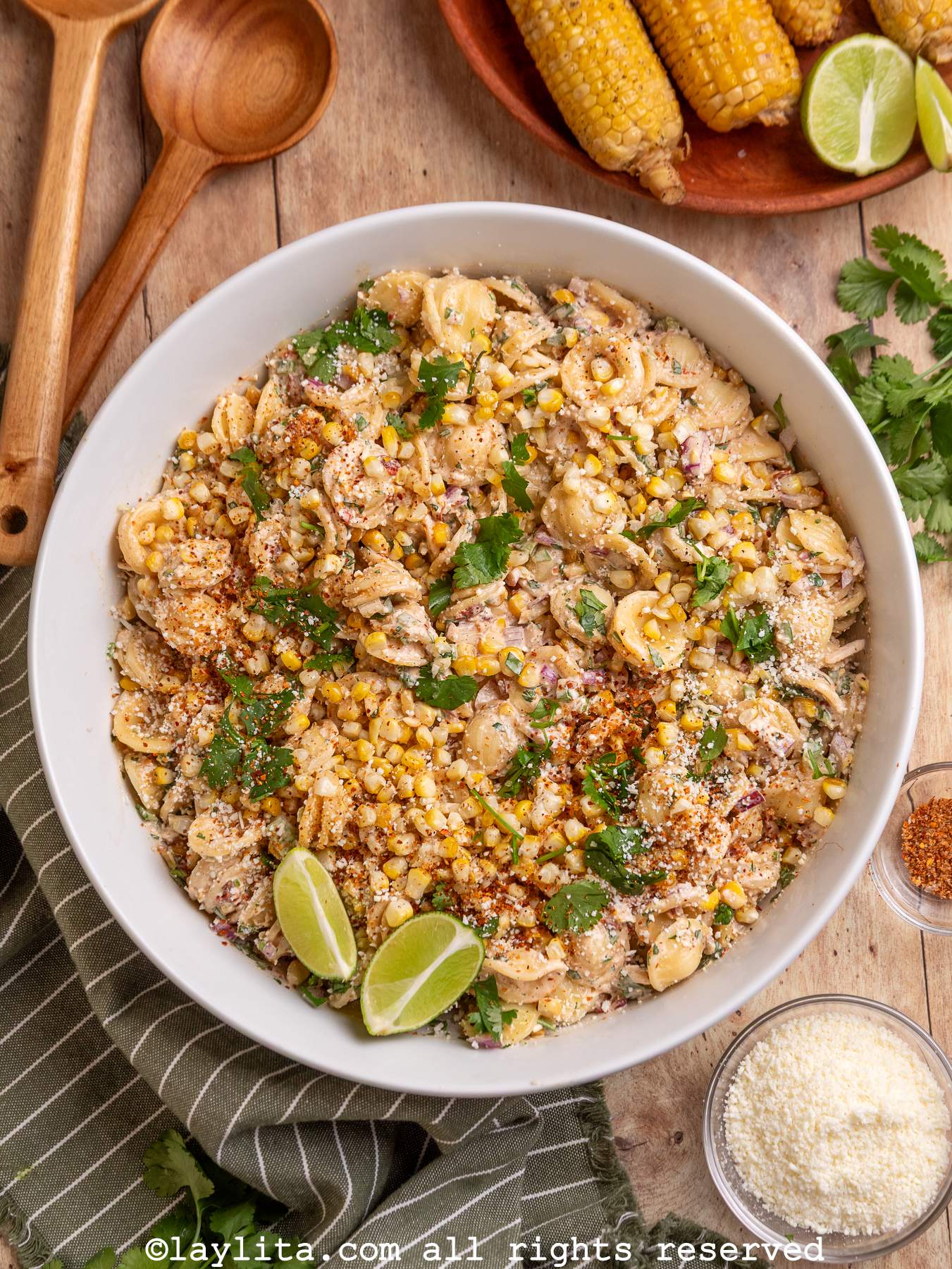 Overhead shot of a bowl filled with Mexican style corn pasta salad next to a tray of grilled corn, some wooden spoons and a bowl of cotija cheese