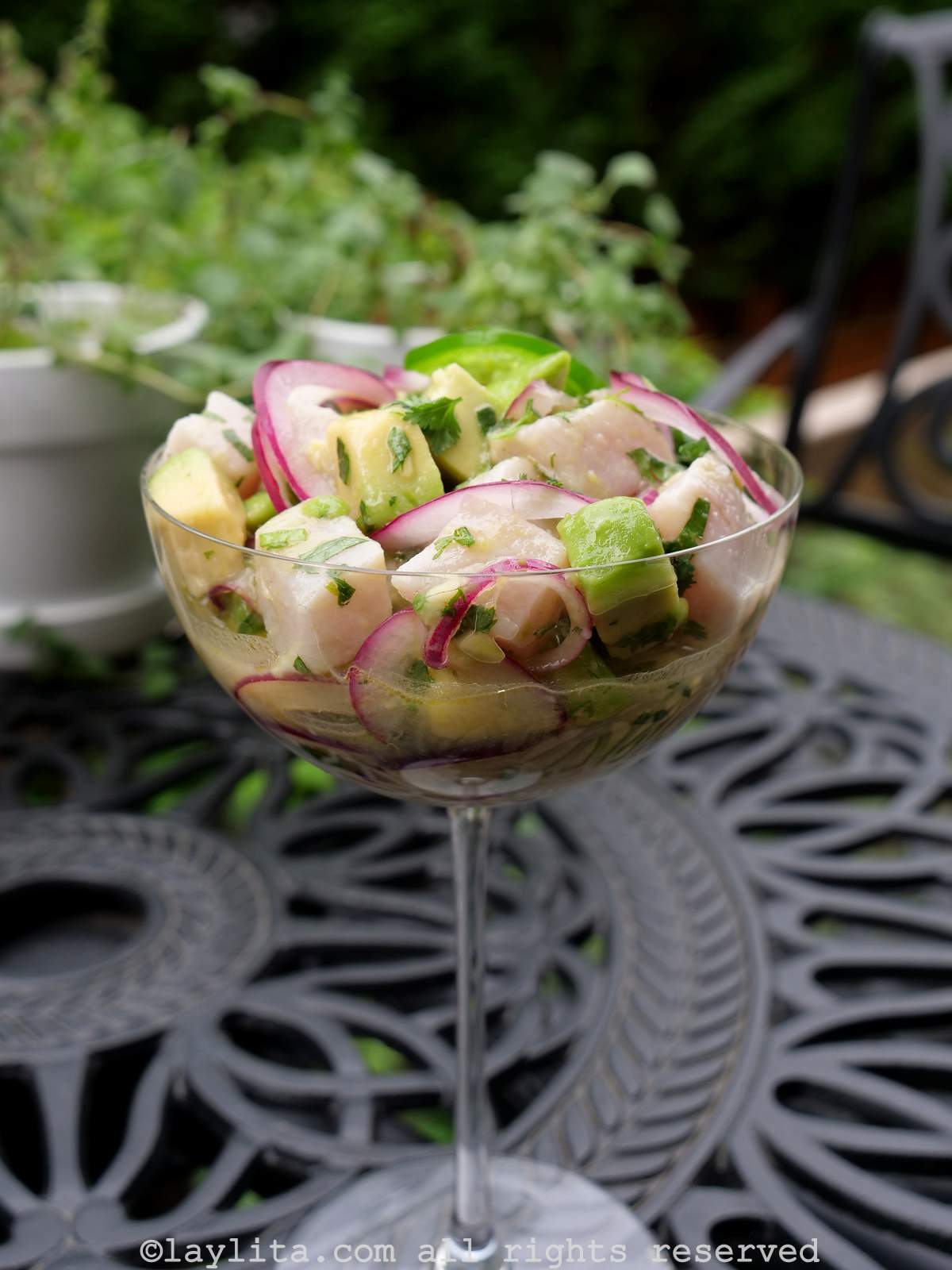 Side view of yellowtail ceviche served in a tall glass on a black garden table with a plant in the background