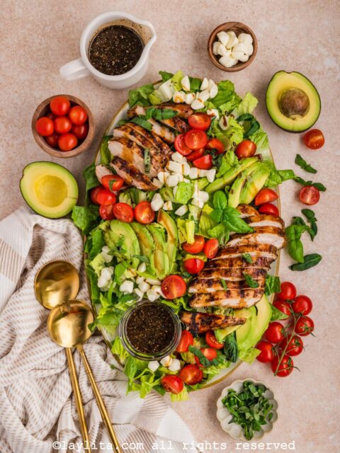Serving platter with a grilled chicken caprese salad with avocados, cherry tomatoes, mozzarella balls, and herbs on the side