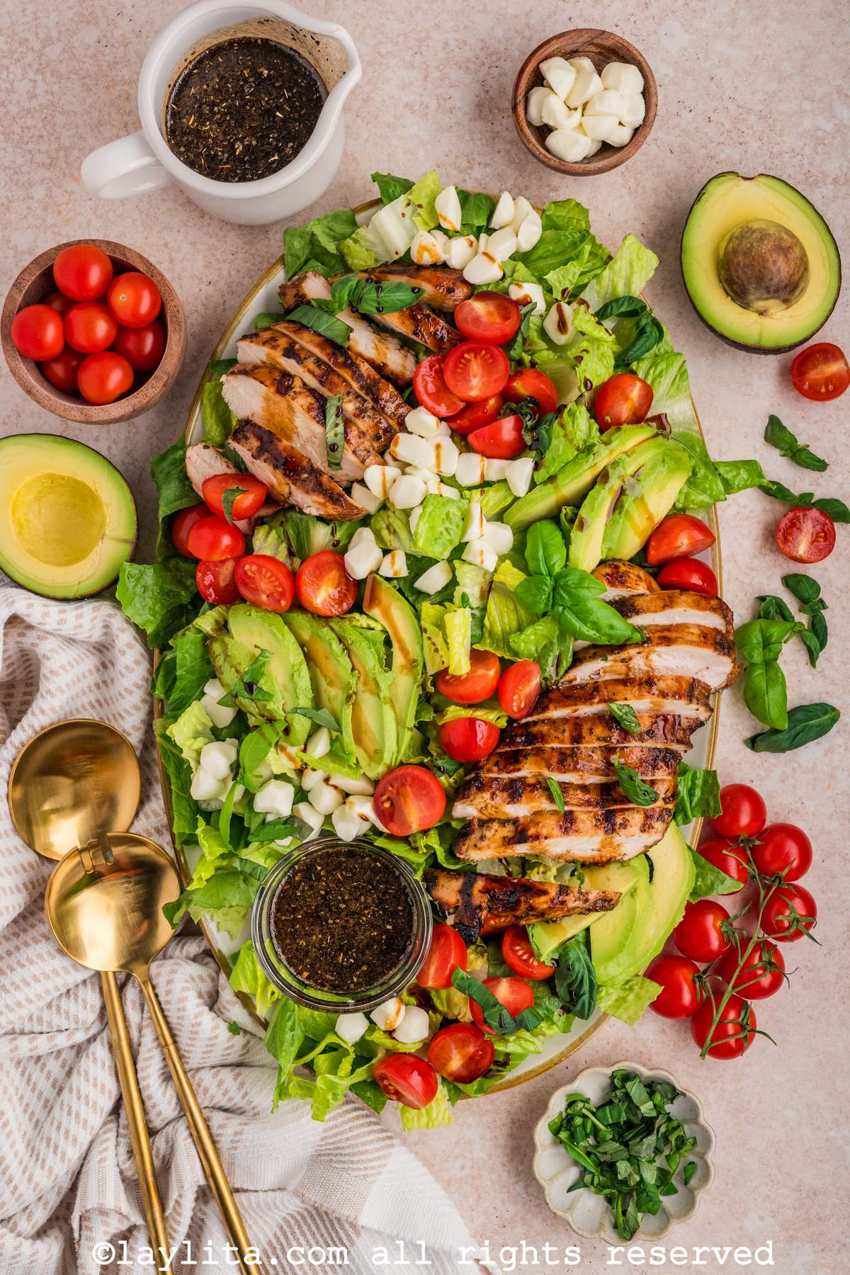 Overhead shot of a large serving platter with a grilled chicken caprese salad with avocados, cherry tomatoes, mozzarella balls, and herbs on the side