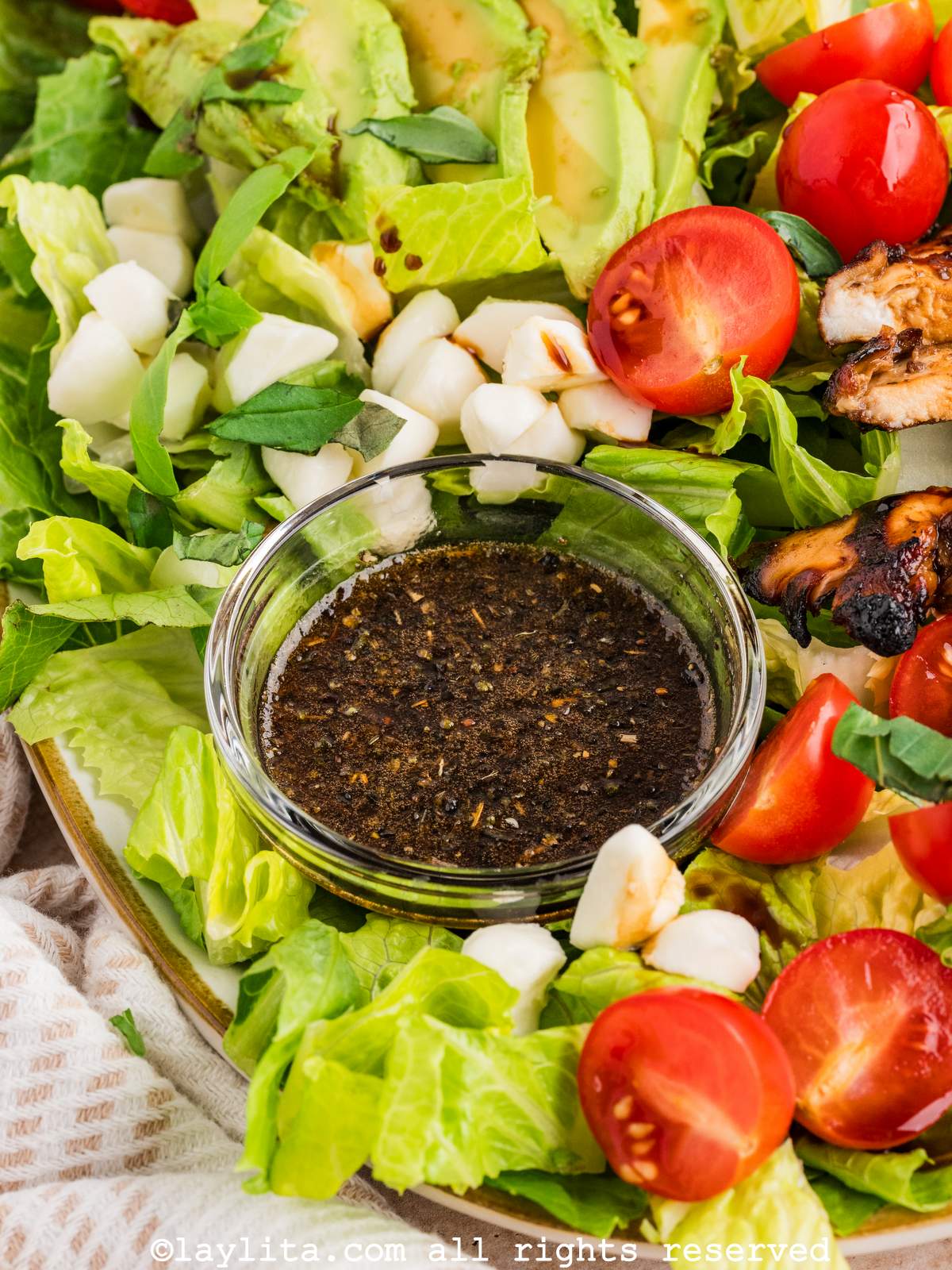 A bowl of balsamic vinaigrette dressing in the middle of a salad platter of lettuce, tomatoes, mozzarella balls and avocado