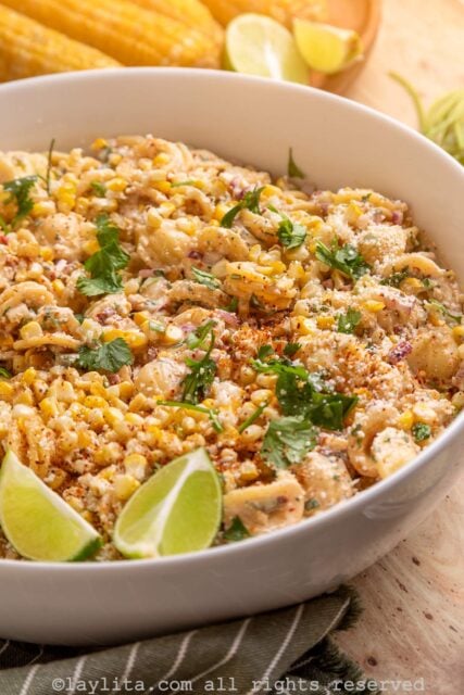 A white bowl filled with corn pasta salad garnished with lime wedges and chopped cilantro leaves