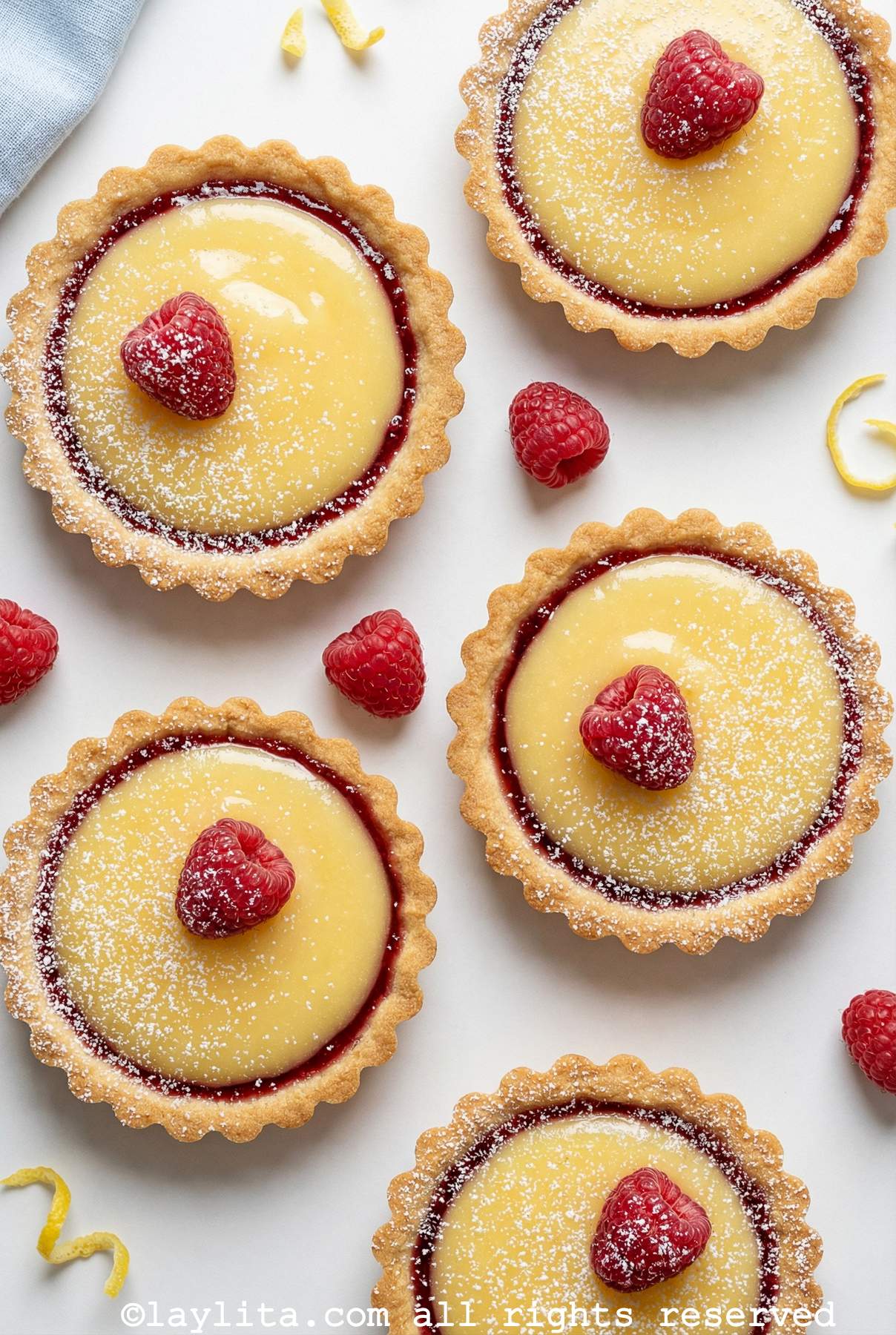 Overhead shot of five raspberry lemon tartlets all with one raspberry on top and some powdered sugar