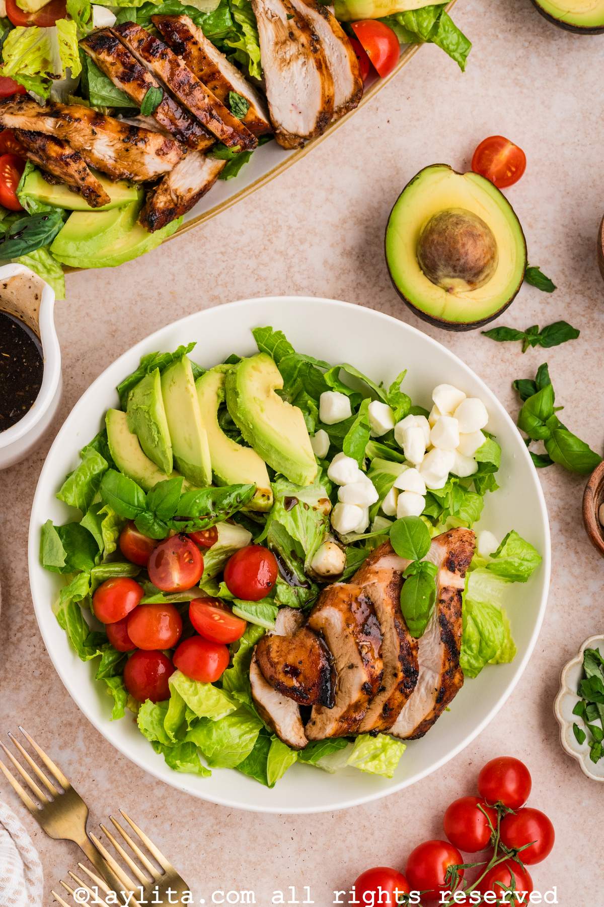 A white bowl filled with lettuce salad and topped with mozzarella, tomatoes, avocado slices and grilled chicken. In the background there is a platter with more salad on it.