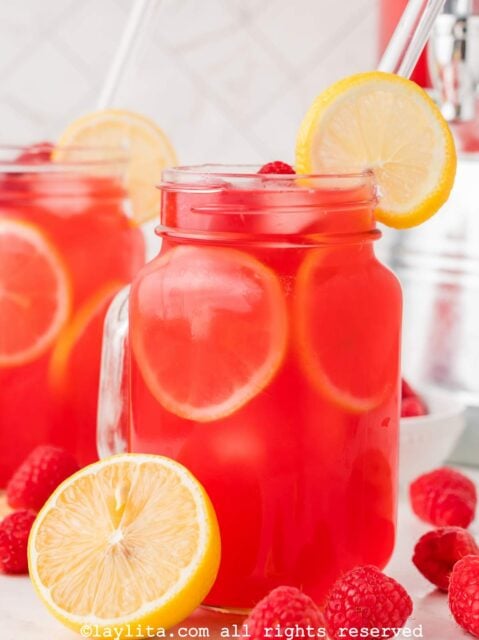 Closeup of a mason jar filled with raspbery lemonade and garnished with a lemon slice and a clear straw, next to a lemon half and more raspberries