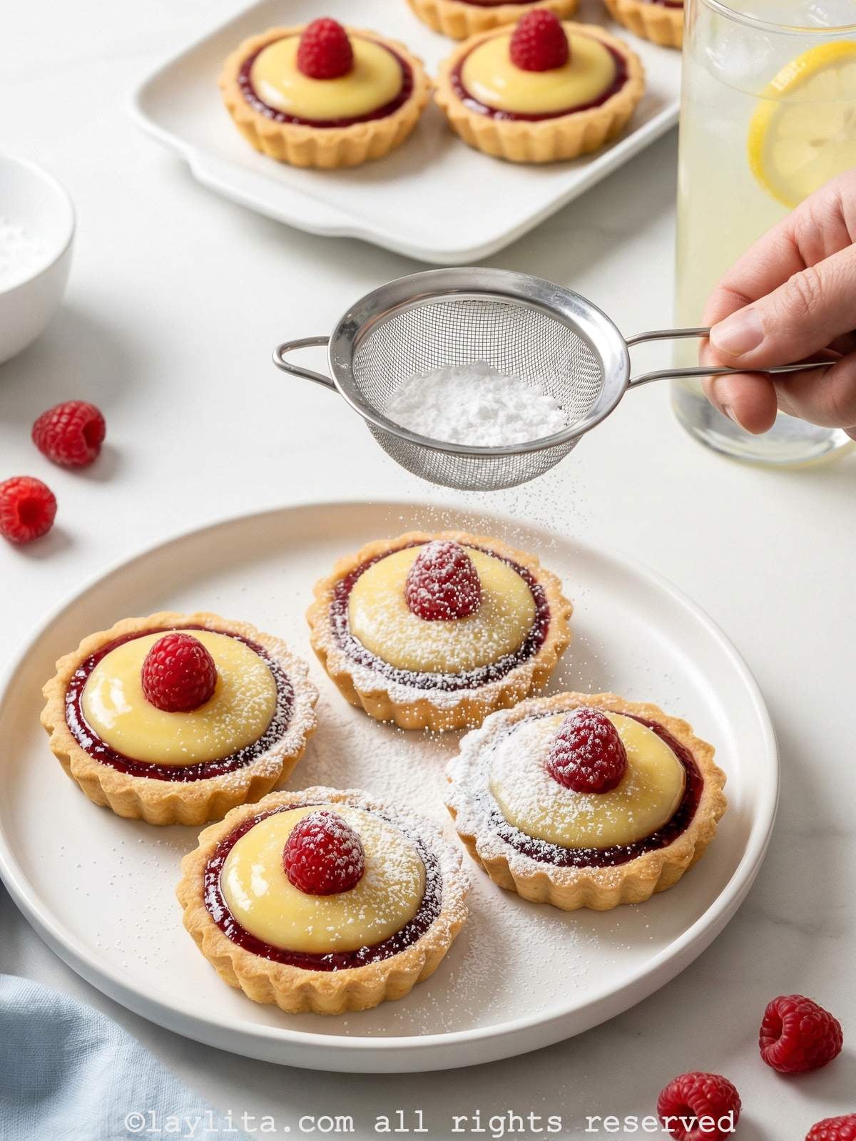 Four lemon raspberry tartlets on a white plate with a hand sprinkling powdered sugar through a strainer on top of them