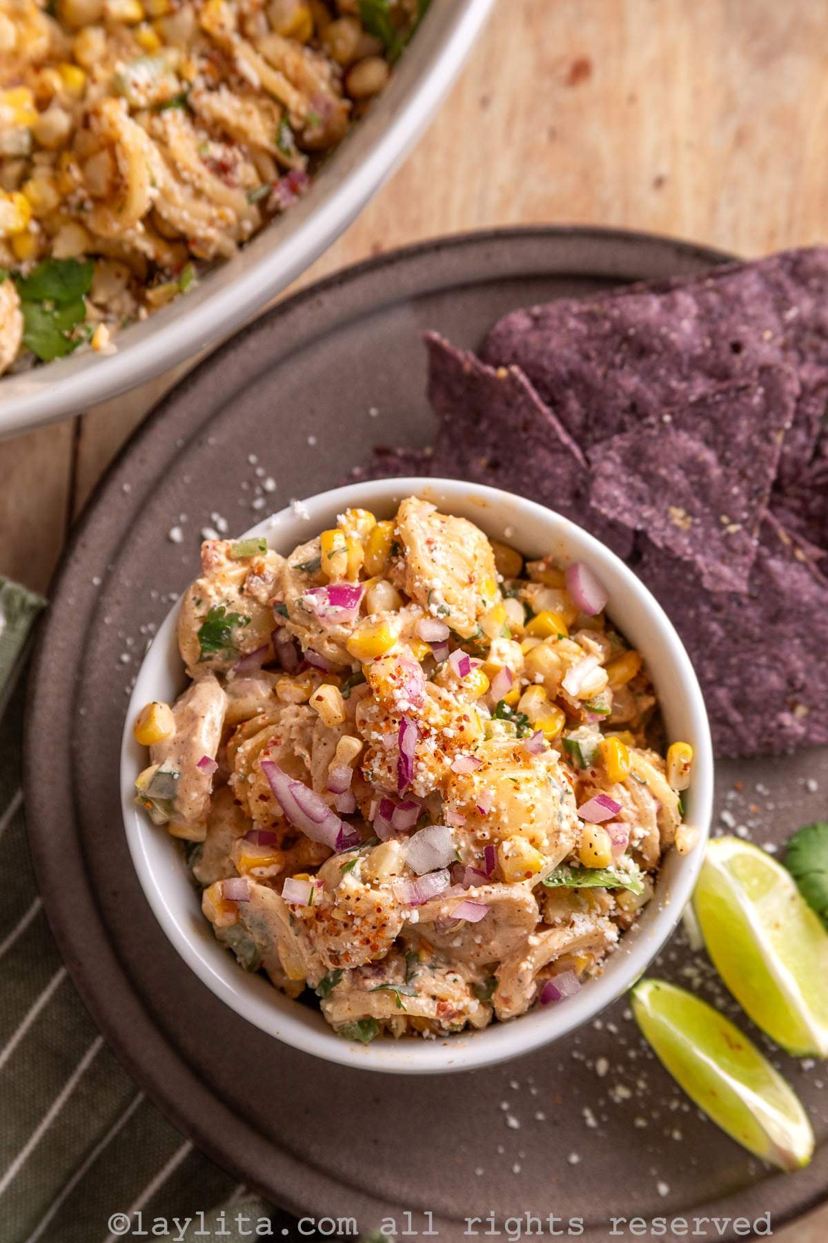A small bowl full of corn pasta salad on a larger platter with purple corn chips and two lime wedges on the side