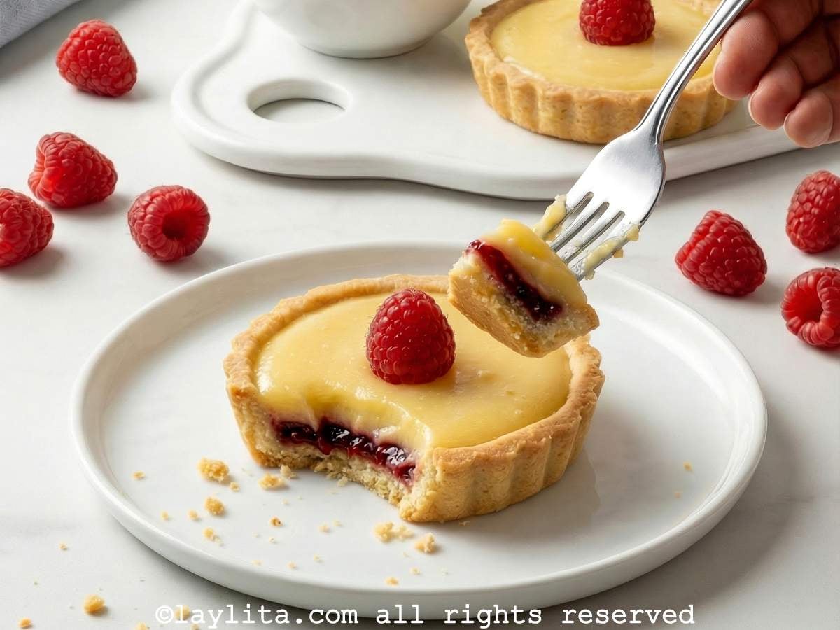 Raspberry lemon tartlet on a plate with a hand holding a fork with a bite of tart on it.
