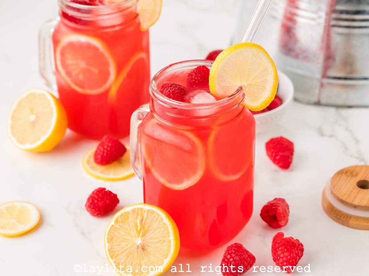 Two mason jar glasses of raspberry lemonade both garnished with lemon slices on a counter with raspberries and lemons spread around.