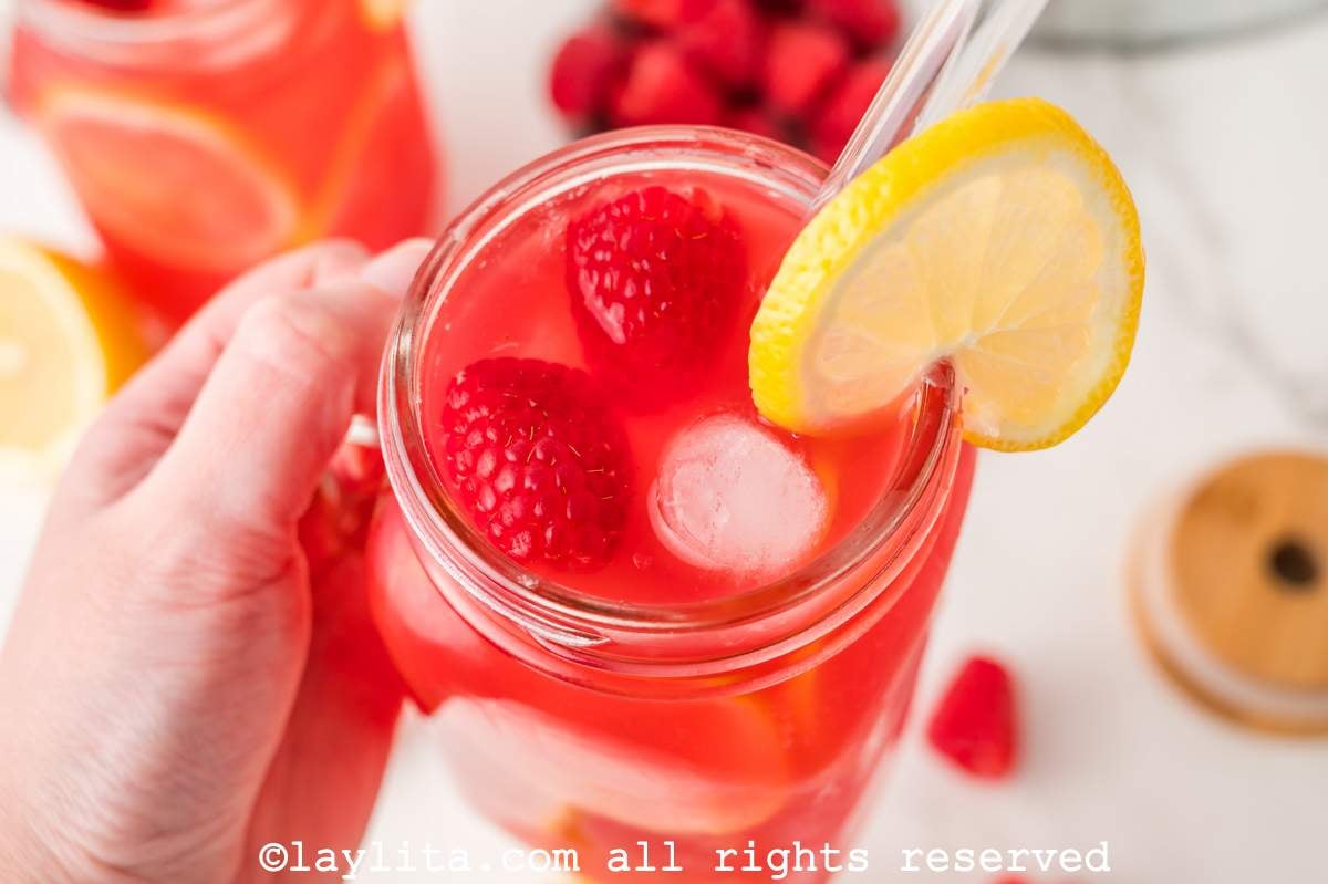 Hand holding a jar of raspberry lemonade, garnished with lemon and fresh raspberries.