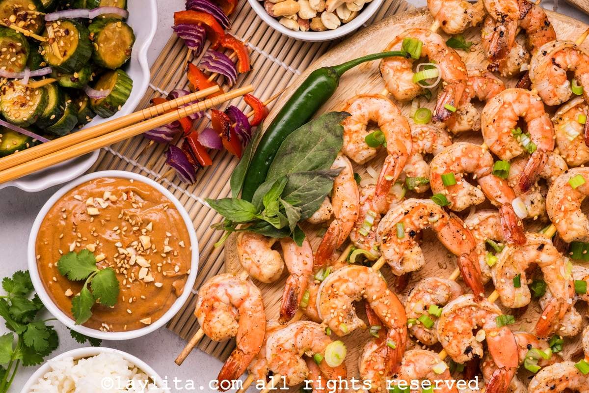 Side view of a platter of grilled shrimp skewers with spicy peppers, veggie skewers, cucumber salad and peanut sauce on the side