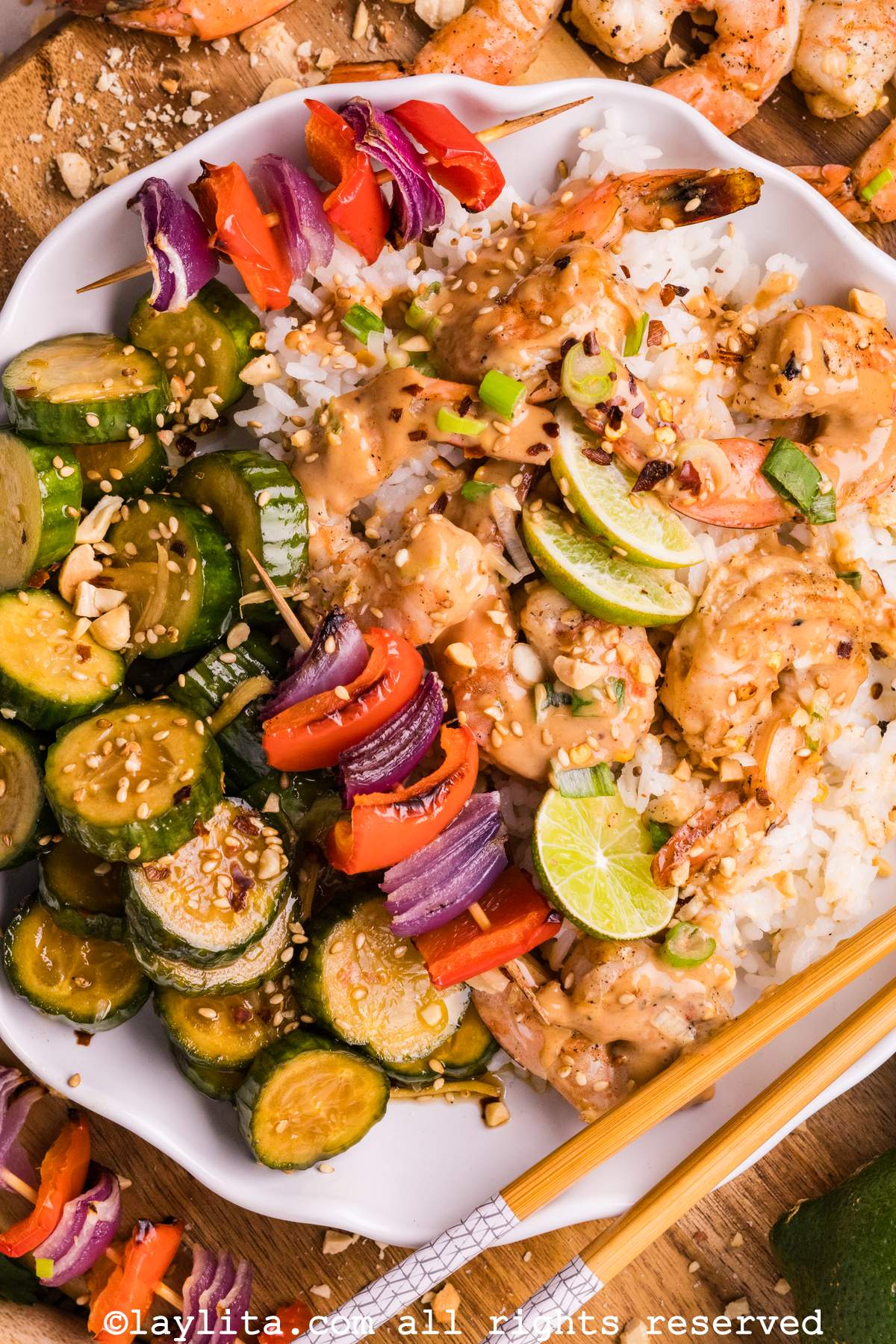 Overhead view of a large white plate with rice, shrimp with peanut sauce, grilled veggies, a cucumber salad and slices of lime