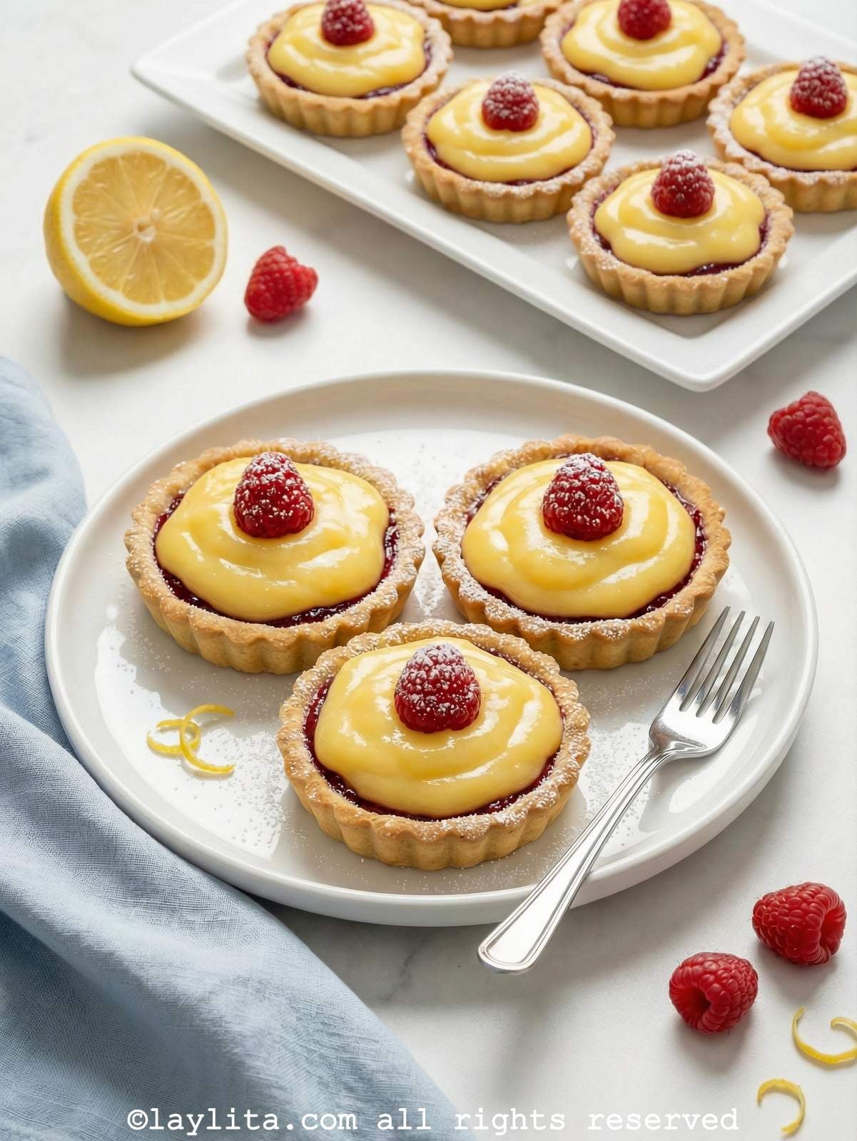 View of three mini raspberry lemon curd tarts on a plate, with several more tarltets on a platter in the bakground