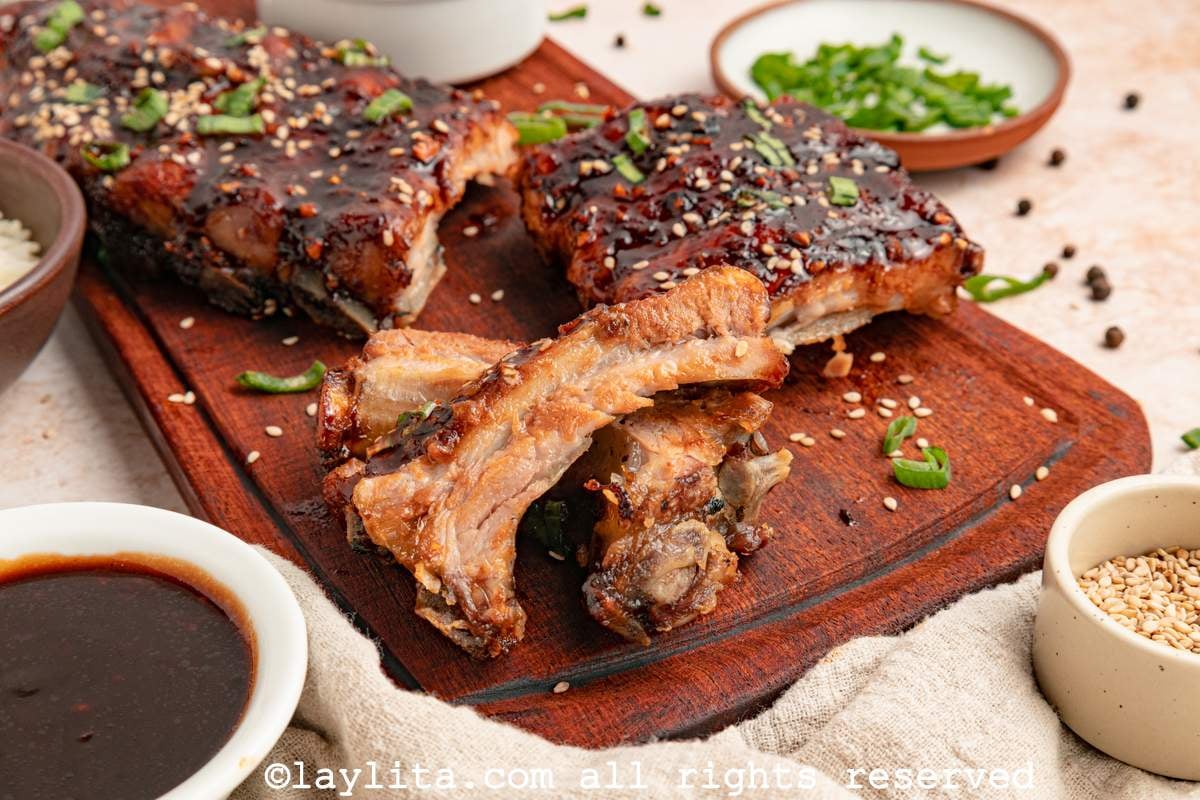 Side view of Asian style ribs on a board, with some cut into slices to show the meat, surrounded by small bowls of sauce, sesame seeds and chopped herbs