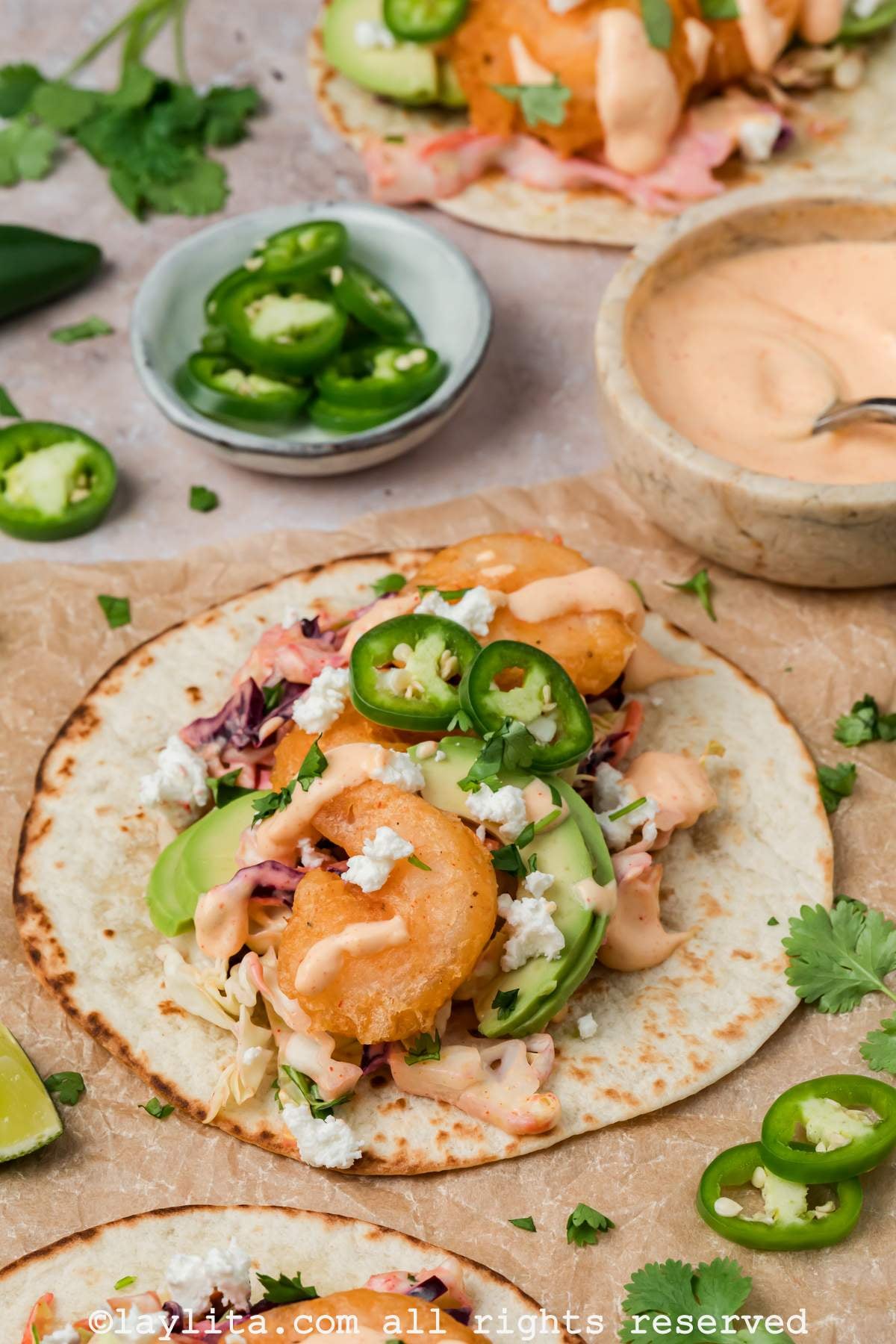 Side view of an open baja shrimp taco on brown parchment paper, with a bowl of sauce and sliced jalapeños in the background
