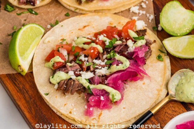 A shredded brisket taco topped with avocado salsa, pickled red onions, pico de gallo and cotija cheese, with lime wedges on the side