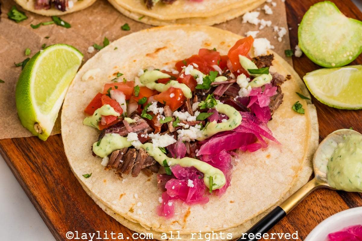 A shredded brisket taco topped with avocado salsa, pickled red onions, pico de gallo and cotija cheese, with lime wedges on the side
