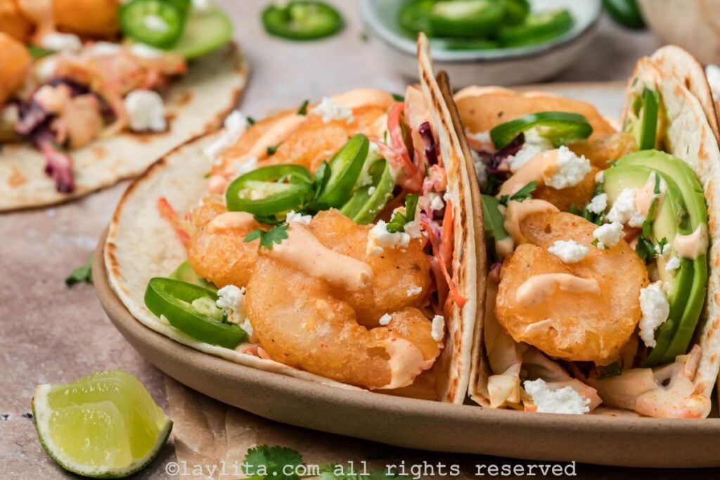 Side view of two baja shrimp tacos on a plate, with crispy shrimp, coleslaw, and baja sauce on top.