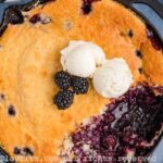 Overhead shot of blackberry cobbler in a cast iron skillet with some missing from the skillet. There are some fresh berries and two scoops of ice cream on top of the cobbler.