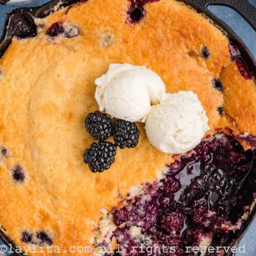 Overhead shot of blackberry cobbler in a cast iron skillet with some missing from the skillet. There are some fresh berries and two scoops of ice cream on top of the cobbler.