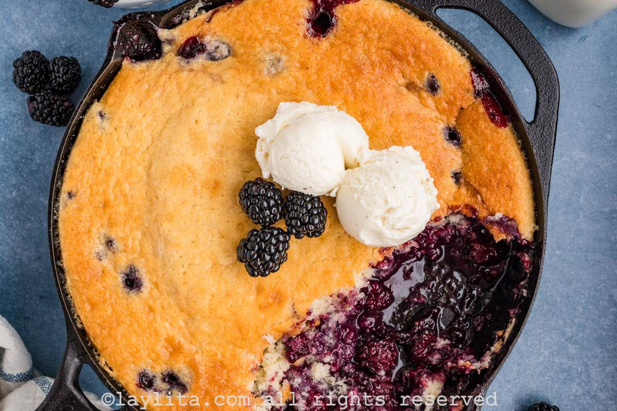 Overhead shot of blackberry cobbler in a cast iron skillet with some missing from the skillet. There are some fresh berries and two scoops of ice cream on top of the cobbler.
