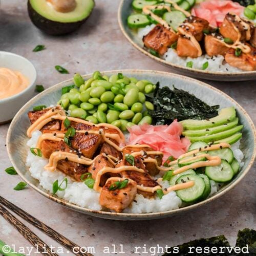 Side view of two salmon sushi bowls with salmon bites, vegetables, and spicy mayo drizzled on top.