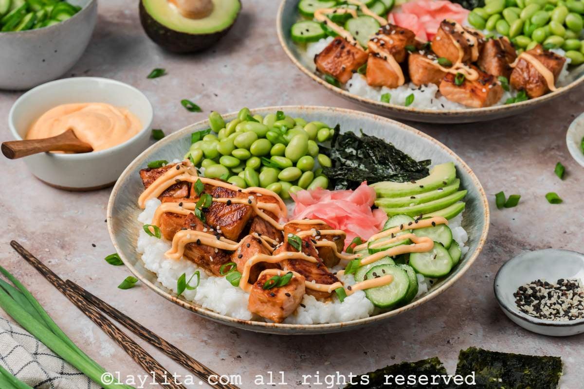 Side view of two salmon sushi bowls with salmon bites, vegetables, and spicy mayo drizzled on top.