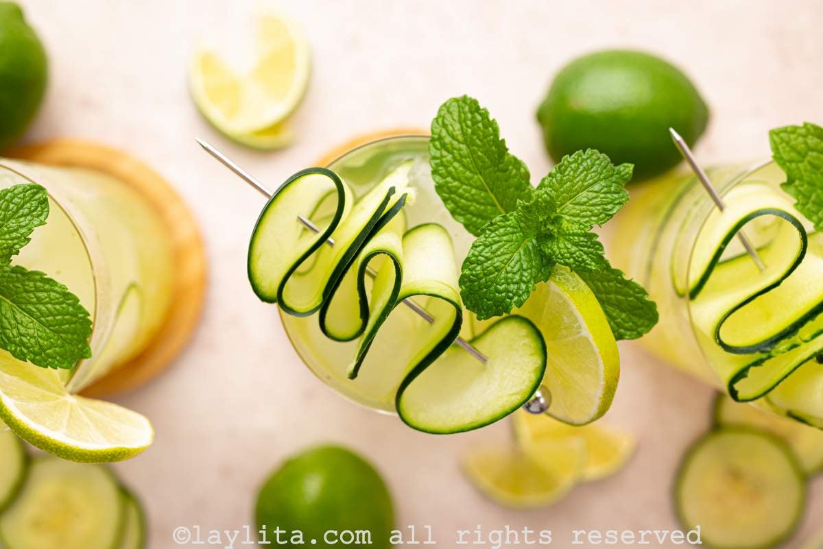 Overhead view of a couple of glasses filled with a cucumber lime drink, garnished with cucumber ribbons and mint, with limes scattered around