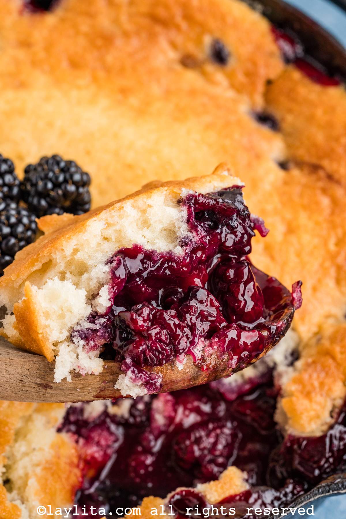 Wooden spoon scooping up some blackberry cobbler from a skillet.