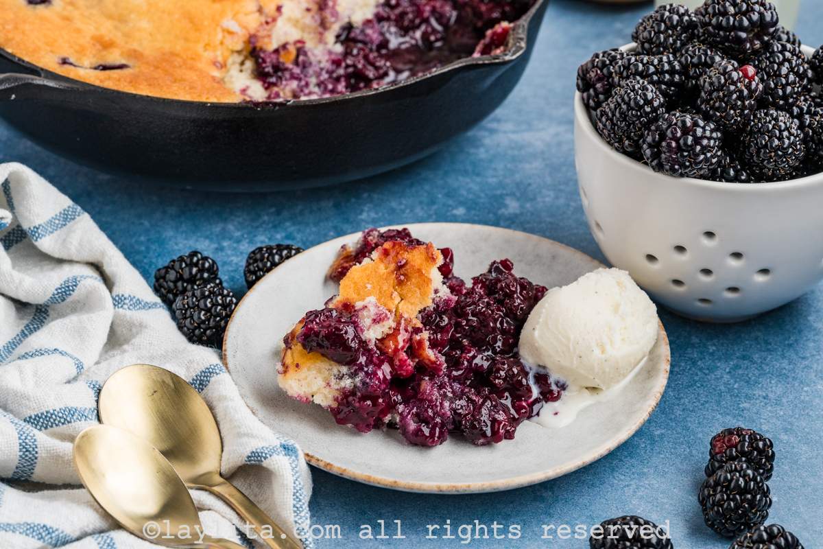 Blackberry cobbler served on a plate with ice cream on the side and the rest of the cobbler and a bowl of berries in the background