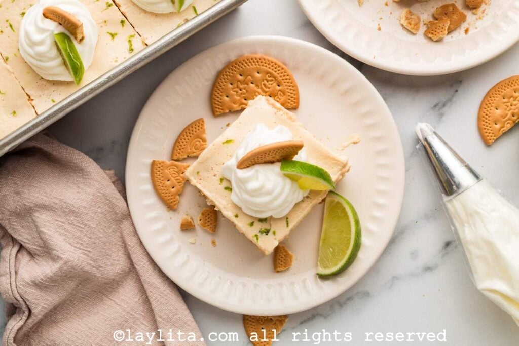 Overhead view of a slice of Mexican lime icebox cake on a plate with cookies and a lime wedge, on the side there is a pastry bag filled with whipped cream