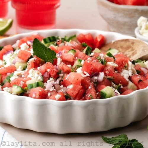 Watermelon cucumber feta salad in a white serving bowl with a serving spoon, around the bowl there are red drinks, lime wedges, mint leaves and a bowl of crumbled feta cheese