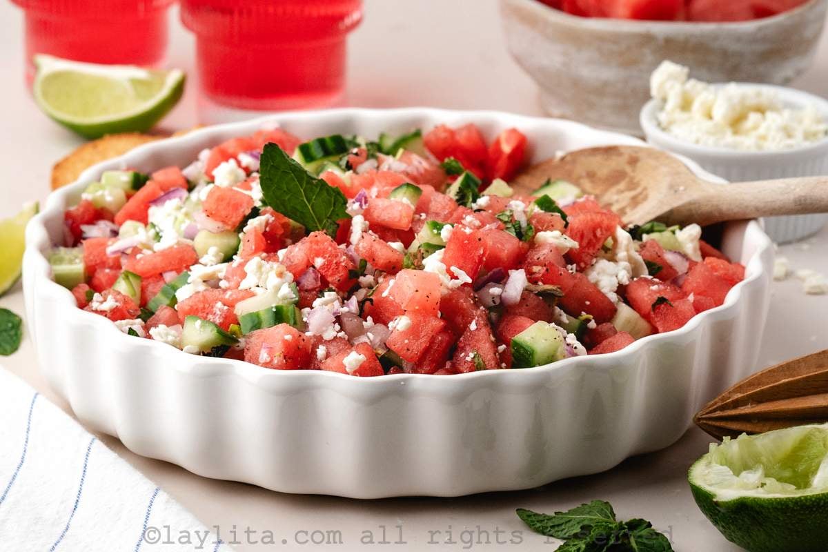 Watermelon cucumber feta salad in a white serving bowl with a serving spoon, around the bowl there are red drinks, lime wedges, mint leaves and a bowl of crumbled feta cheese