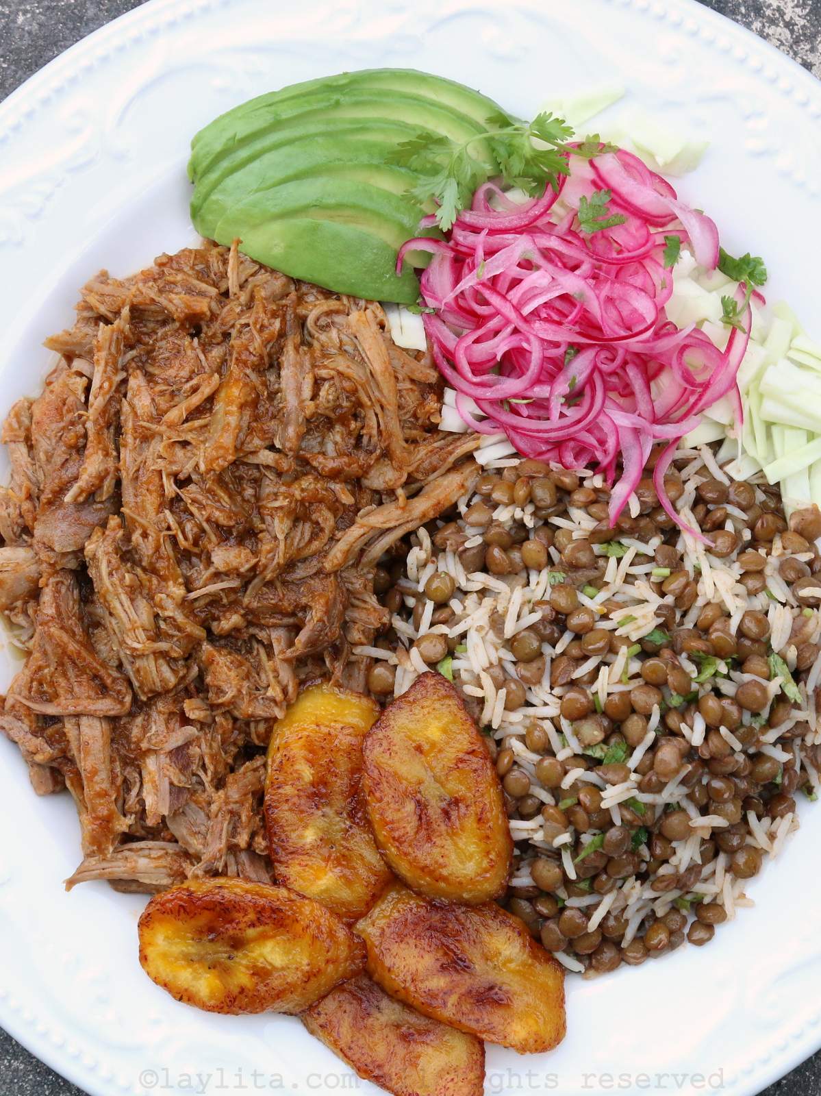 Overhead shot of chicha pulled pork served with rice and lentils, fried plantains, avocado and pickled onions on a plate.
